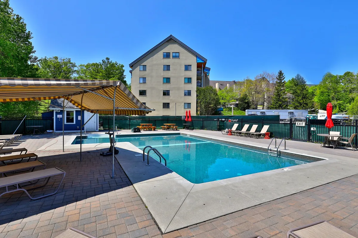 An inviting exterior view of the resort's swimming pool, surrounded by loungers and an awning, with green trees and a building visible in the background. The area is set up for leisure, socializing, and relaxation, hinting at the outdoor amenities available to guests.