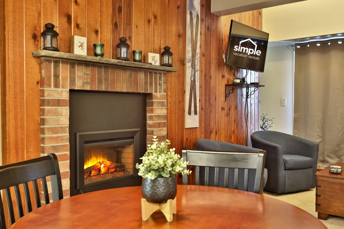 The image showcases a cozy studio interior featuring a stone fireplace as the focal point, with a comfortable seating area, a dining table set for four, a television, and tasteful alpine-themed décor. The scene is warm and inviting, suggesting a perfect spot for relaxing after a day on the slopes or sharing meals with loved ones.