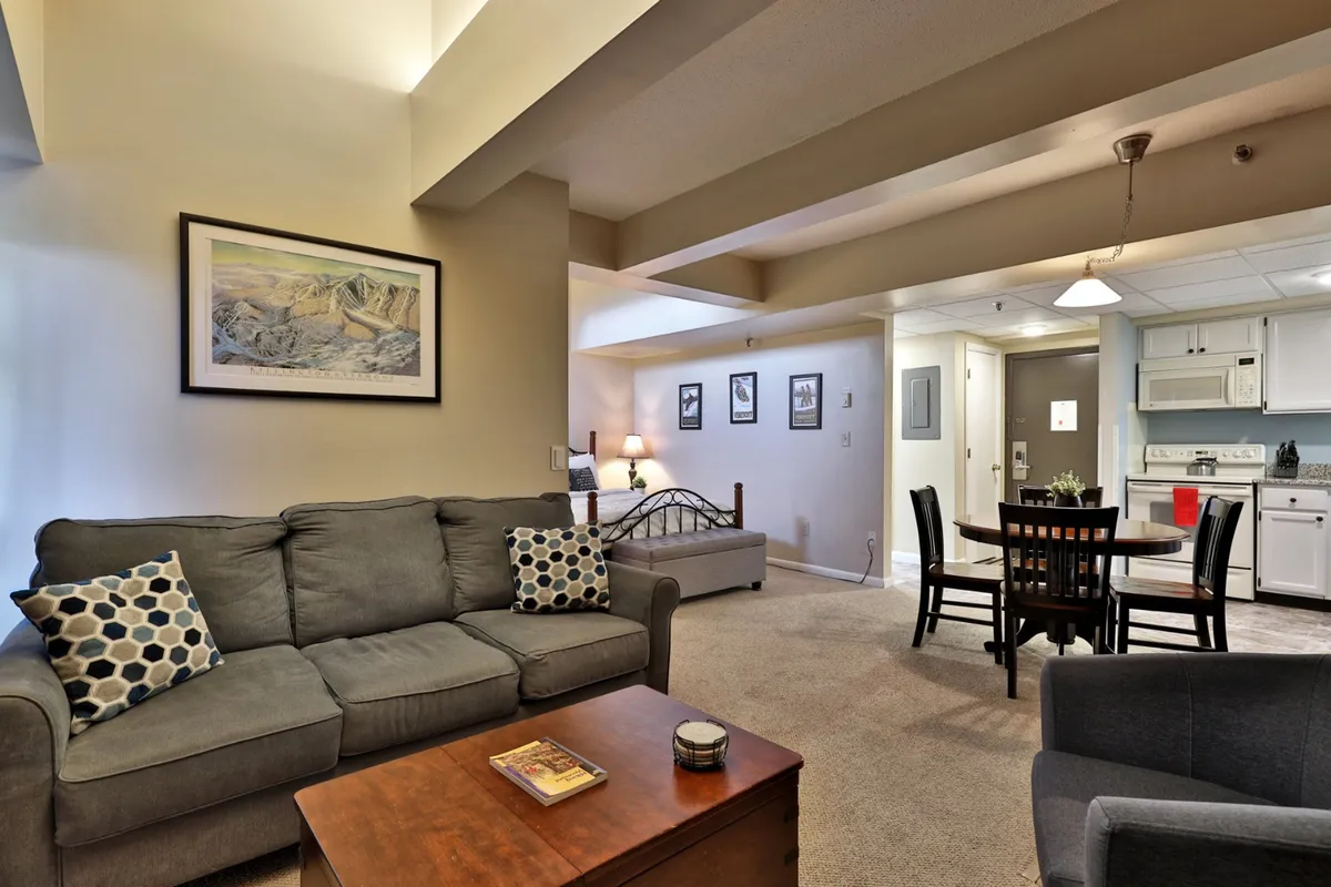 This cozy open-layout living area features a sofa with decorative pillows, a wooden coffee table, a dining table with chairs, and a visible kitchen area. The space is styled for warmth and togetherness, suggesting family meals, relaxing evenings, and time spent together in comfort.