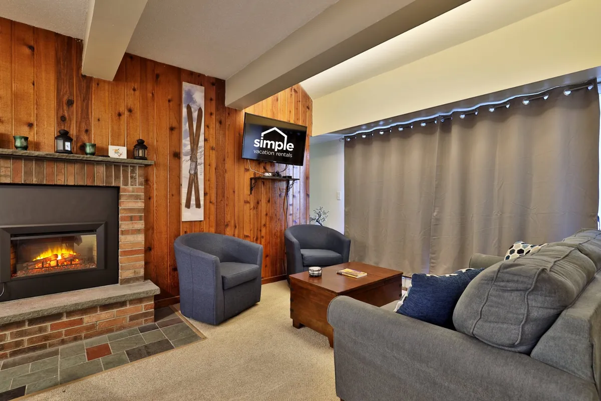 The image features a warm seating area centered on a glowing fireplace, with wood paneling and a television mounted nearby. Plush seating invites guests to relax and enjoy the ambiance, ideal for unwinding after a day outdoors or watching a favorite show.