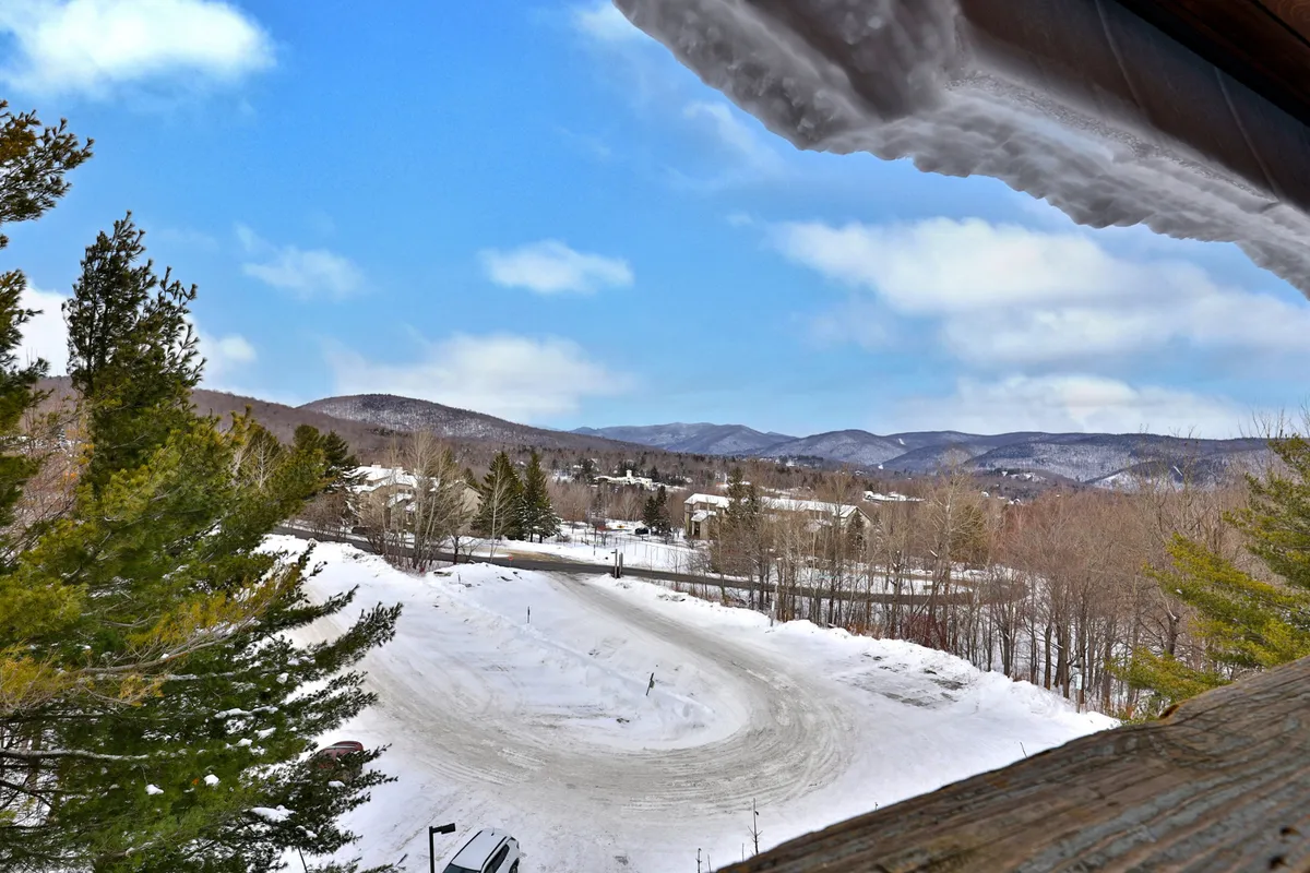 A sweeping winter scene featuring snow-covered mountains, a winding road framed by trees laden with fresh snow, and residential buildings in the foreground. The tranquil landscape is ideal for winter sports enthusiasts and showcases the proximity to both the slopes and local amenities.