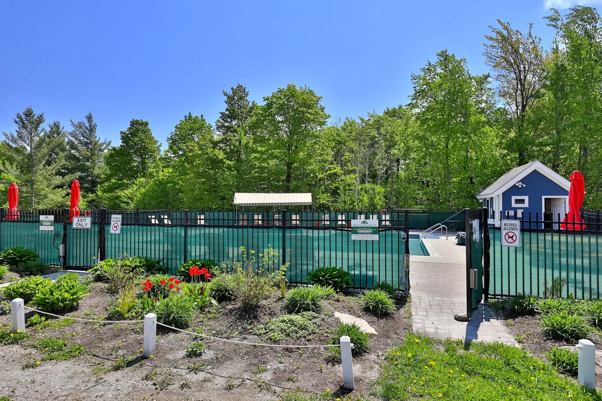 A cheerful exterior scene featuring the resort's swimming pool, surrounded by lush greenery, red umbrellas, garden flowers, and a charming pool house. The area is lively yet relaxing, emphasizing communal enjoyment and resort amenities in a garden-like setting.